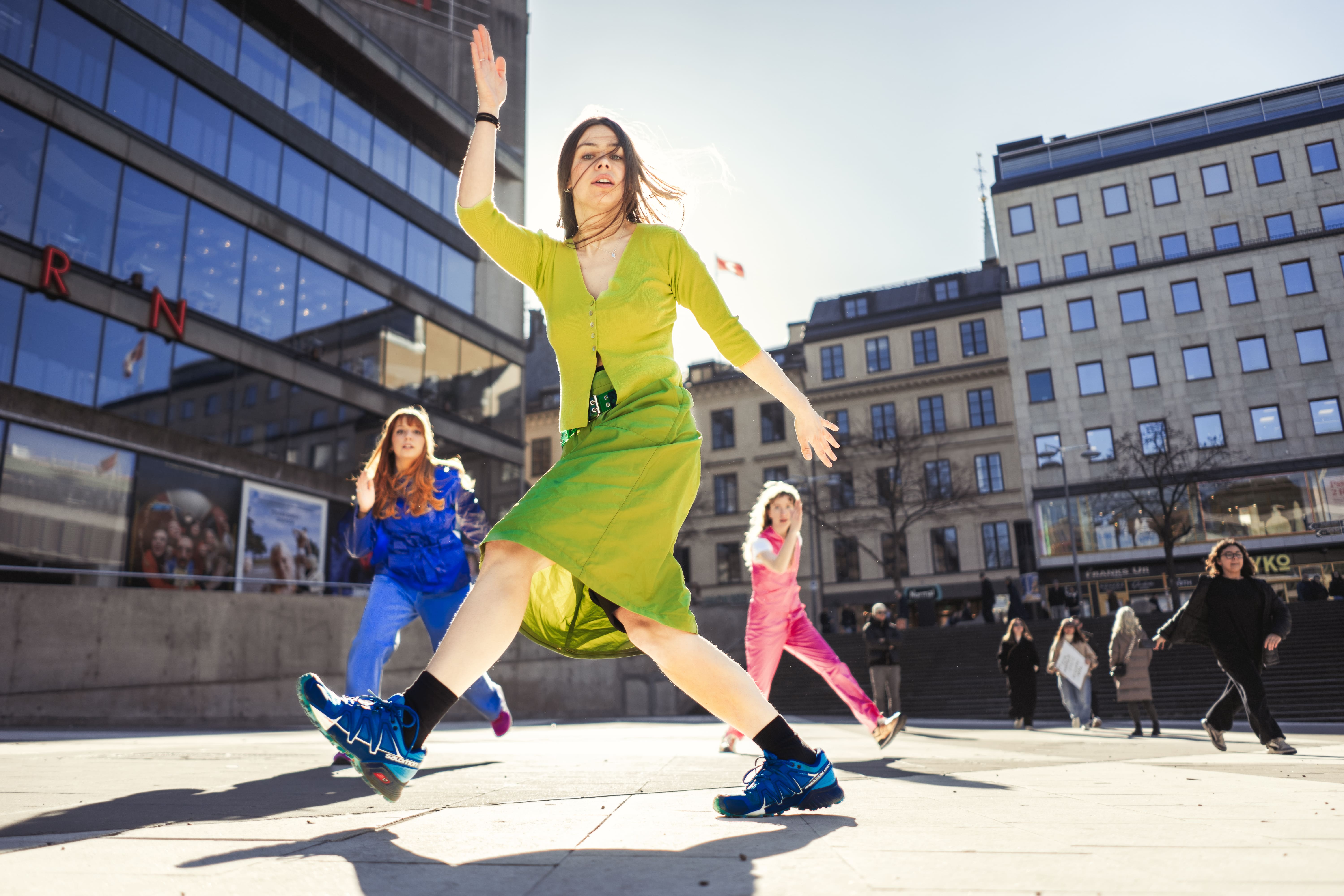 Tre dansare på ett torg i grönt, rosa och lila  
