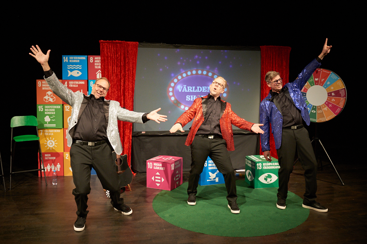 Tre herrar i glitterkavajer dansar framför scenografi med Globala målen