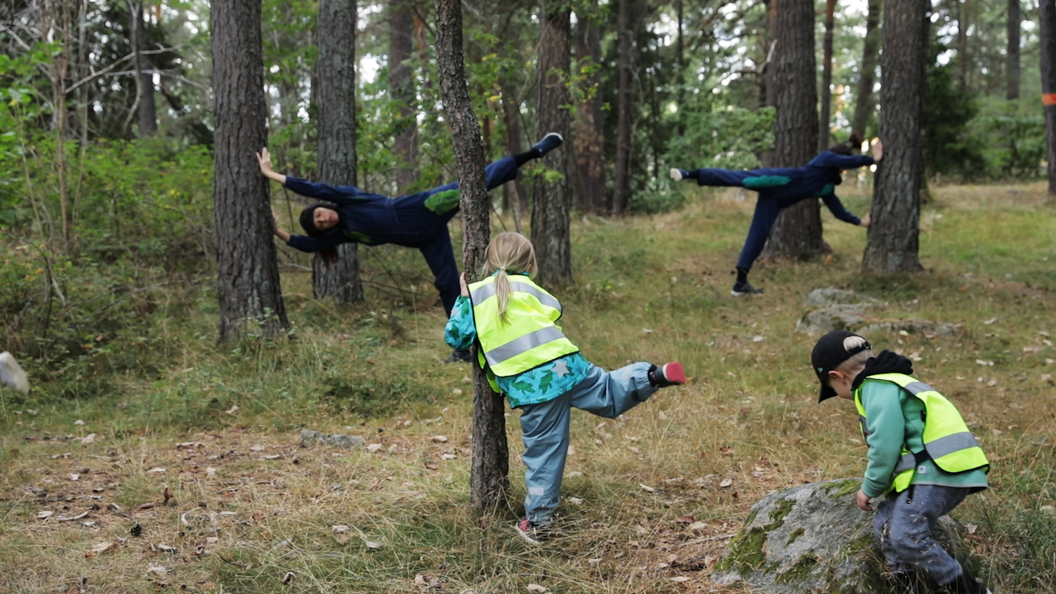 I skogen bland träden. Två dansare lutar mot trädstammar med sina händer, står på ett ben, det andra benet ut. Barnen dansar med.