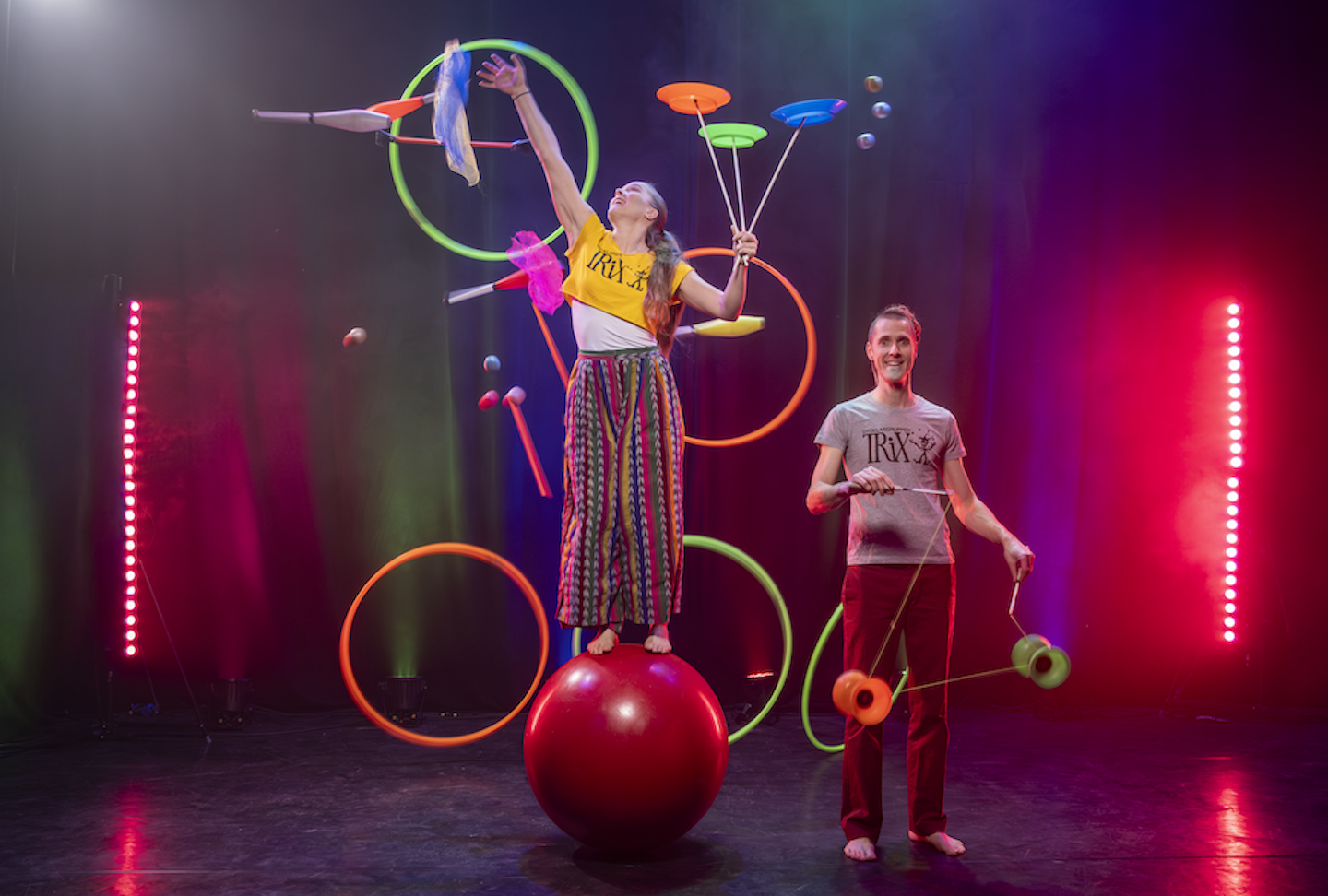 En artist ballanserar på ett cirkusklot och en annan artist står bredvid. Båda jonglerar.